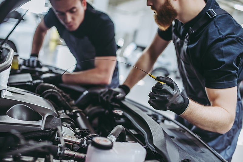 Two mechanics inspecting a car engine, one checking the oil with a dipstick