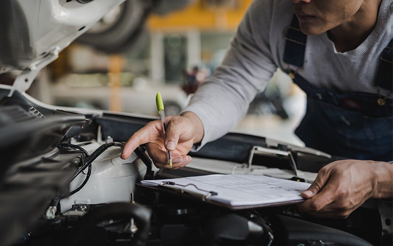 Technician inspecting a car engine and writing notes on a clipboard