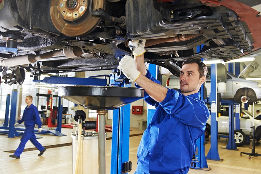 Auto mechanic in blue uniform working underneath a raised car in a repair shop.
